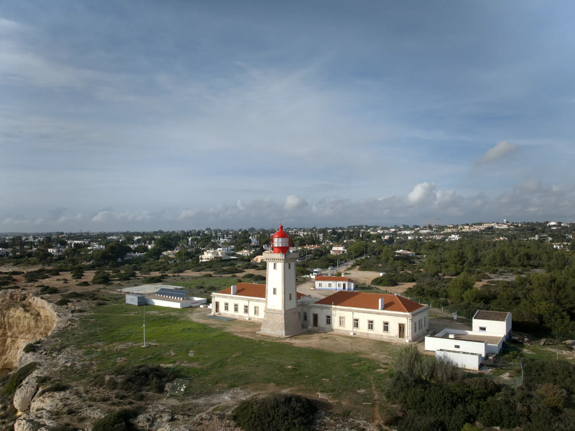 Farol de Alfanzina seen from the air: a square white lighthouse tower with a red lantern on the cliff top above the sea